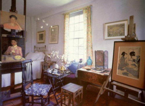 A cozy artist's studio with paintings, a bed, and various furniture, illuminated by natural light from a window.