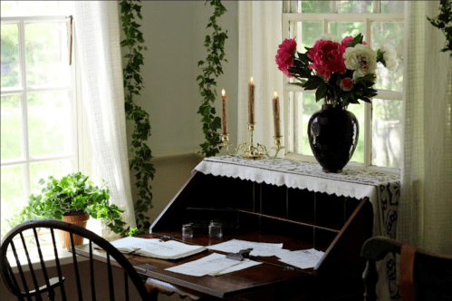 A cozy desk with papers, a vase of flowers, and candles, set by a window with greenery and soft natural light.