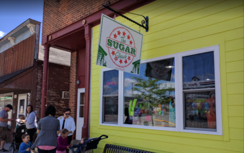 A colorful storefront of "The Sugar Market" with people outside, enjoying a sunny day.