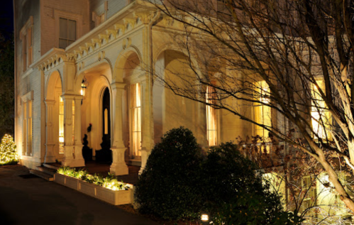 A beautifully lit historic mansion at night, featuring arched doorways and lush greenery.