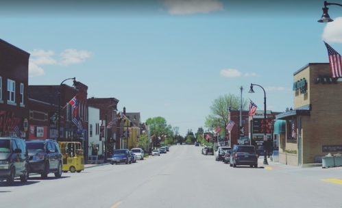 A sunny street view of a small town lined with shops and American flags, featuring parked cars and clear blue skies.