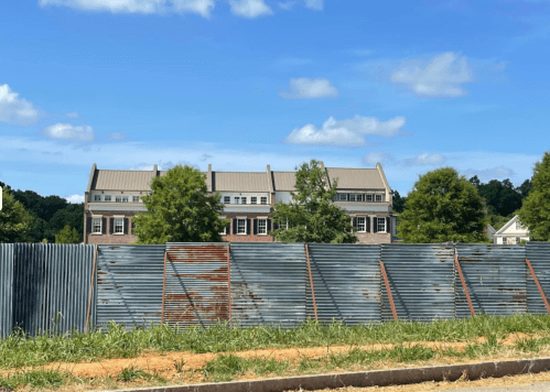 A corrugated metal fence in the foreground with a building and trees visible in the background under a blue sky.