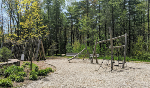 A sunny playground with wooden swings and climbing structures, surrounded by trees and gravel pathways.
