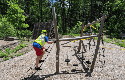 A person in a bright vest climbs a wooden playground structure in a wooded park area.
