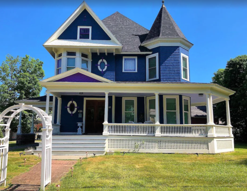 A large, blue Victorian-style house with a wraparound porch and a white fence, set against a clear blue sky.