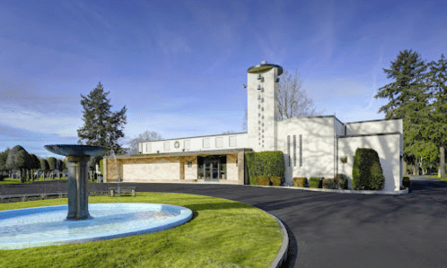 Modern building with a fountain in front, surrounded by manicured lawns and trees under a clear blue sky.