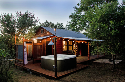 Cozy cabin with string lights and a hot tub on a wooden deck, surrounded by trees in a serene outdoor setting.