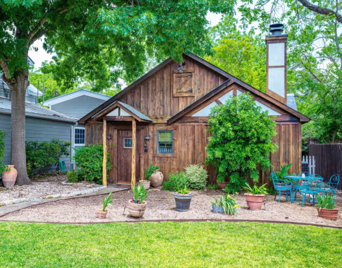 A charming wooden house surrounded by greenery and potted plants, with a small patio and outdoor seating area.
