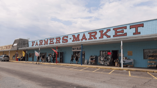 A blue building with "FARMERS MARKET" sign, featuring shops and people walking outside on a cloudy day.