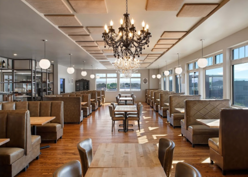 Modern restaurant interior with elegant booths, wooden tables, and a chandelier, featuring large windows and a scenic view.