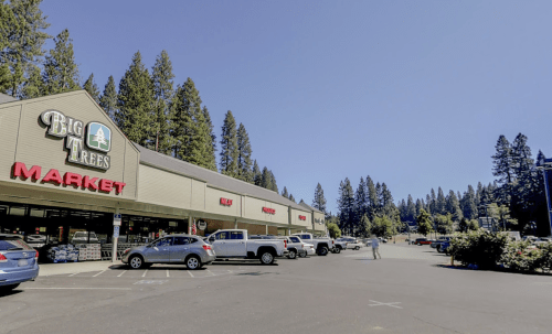 A grocery store named "Big Trees Market" with a parking lot surrounded by tall trees under a clear blue sky.