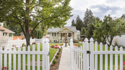 A charming house with a white picket fence, surrounded by a lush garden and colorful flowers under a cloudy sky.
