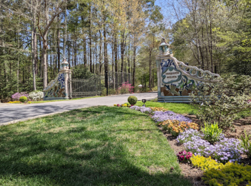 A decorative entrance gate surrounded by colorful flowers and greenery, leading into a wooded area.