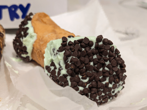 A close-up of a cannoli filled with mint green cream and coated in mini chocolate chips.