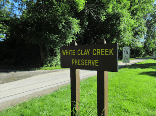 Sign for White Clay Creek Preserve beside a road, surrounded by greenery and trees. Speed limit sign visible in background.