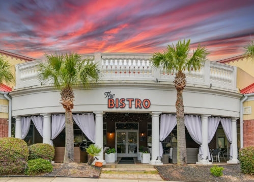 A charming bistro with palm trees, draped curtains, and a colorful sunset sky in the background.