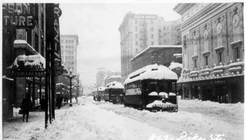 Historic black-and-white photo of a snowy street with streetcars and buildings, capturing a winter scene.