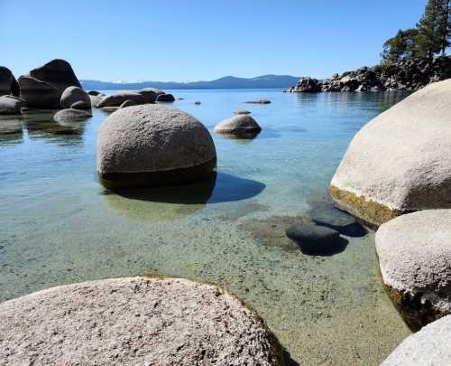 A serene lake scene with smooth, round rocks in clear water under a blue sky and distant mountains.