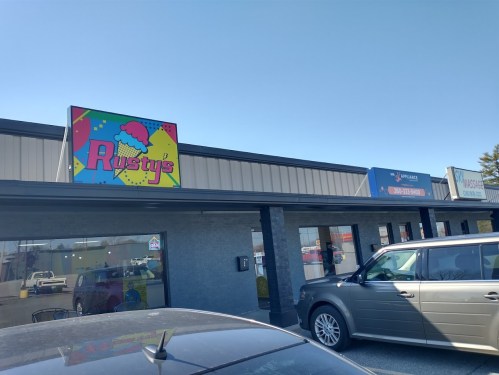 A storefront with a colorful sign reading "Rusty's" and other business signs, parked cars in the foreground.