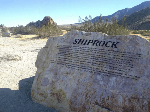 A large rock formation with an inscription about Shiprock, surrounded by desert landscape and mountains in the background.