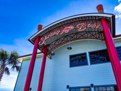 Sign for "Shrimp Boat" restaurant under a blue sky, featuring red pillars and a modern architectural design.