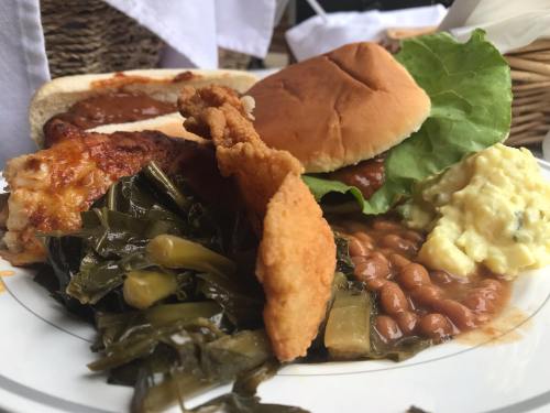 A plate of Southern comfort food featuring fried chicken, collard greens, baked beans, and two sandwiches.