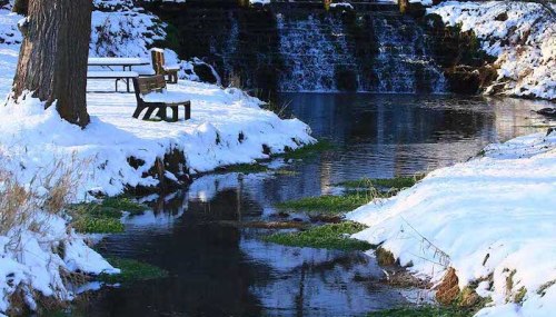 A serene winter scene featuring a snowy landscape, a small creek, and a bench near a gentle waterfall.