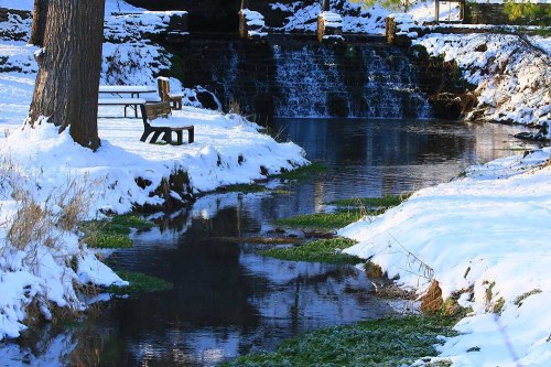 A serene winter scene with a snowy landscape, a small creek, and a bench near a gentle waterfall.