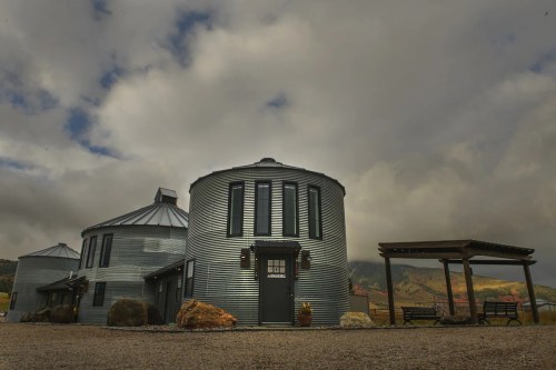 A modern building made from silos, surrounded by rocks and a wooden pergola, under a cloudy sky.