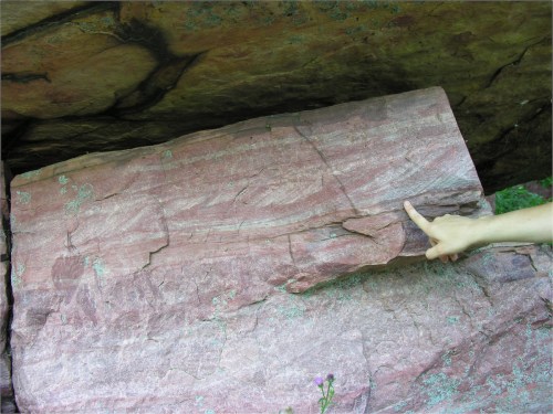 A hand points to a large, layered rock formation with reddish hues and visible textures in a natural setting.