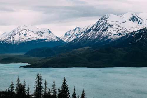 A scenic view of snow-capped mountains surrounding a calm, blue lake under a cloudy sky.