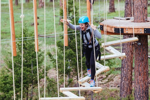 A woman in a blue helmet navigates a rope course, smiling as she steps on wooden planks suspended in the air.