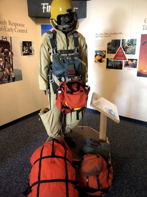 A mannequin in a yellow helmet and protective suit, equipped with backpacks and gear, displayed in an educational exhibit.