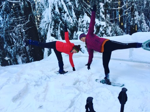 Two people in winter gear balancing on one leg in a snowy forest, surrounded by tall trees.