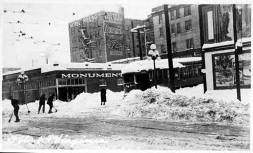 Historic black-and-white photo of a snowy street with people shoveling snow near buildings and a train.