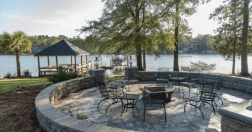 A lakeside patio with a fire pit and seating, surrounded by trees and a gazebo in the background.