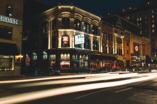 A historic building illuminated at night, with light trails from passing cars and nearby city lights.