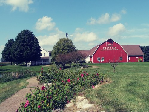 A red barn and white building on a sunny day, surrounded by green grass and blooming flowers along a stone path.