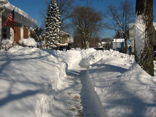 A snowy neighborhood scene with tall snowbanks lining a cleared sidewalk under a clear blue sky.