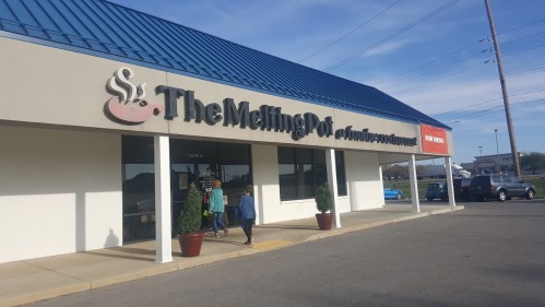 Exterior of The Melting Pot restaurant, featuring a blue roof and large sign, with people entering the building.