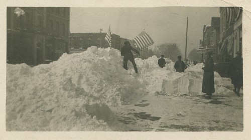 Historic black-and-white photo of people navigating snowdrifts, with flags, in a town street after a heavy snowfall.