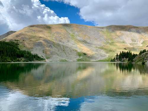 A serene lake reflecting mountains and clouds, surrounded by lush greenery and rocky terrain.