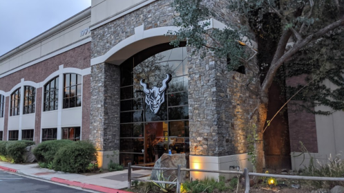 Exterior of a modern building with stone accents and a large bull logo on the glass entrance, surrounded by greenery.