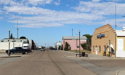 A quiet street scene with buildings on either side, including a pink structure and a tan building, under a blue sky.