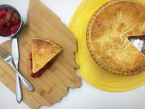 A slice of pie on a wooden board next to a whole pie on a yellow placemat, with a bowl of fruit on the side.