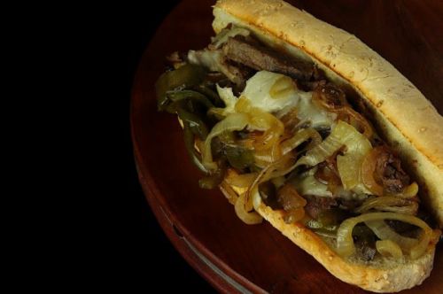 A close-up of a sandwich filled with sliced beef, melted cheese, sautéed onions, and green peppers on a toasted roll.