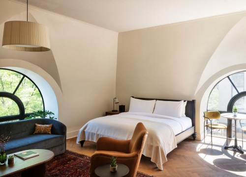 A modern hotel room featuring a large bed, stylish furniture, and arched windows with greenery outside.