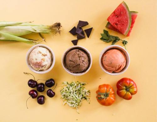 Three bowls of ice cream on a yellow background, surrounded by fresh fruits and vegetables like cherries, watermelon, and corn.