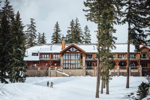 A snowy landscape featuring a large wooden lodge surrounded by tall pine trees and two people walking in the snow.