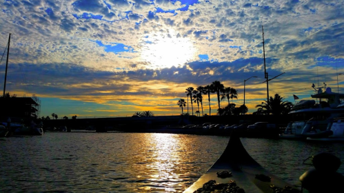 A serene marina at sunset, with vibrant clouds reflecting on the water and palm trees silhouetted against the sky.